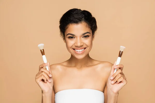 Joyeuse femme afro-américaine aux épaules nues tenant des pinceaux cosmétiques isolés sur beige — Photo de stock