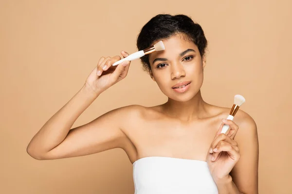 Jeune femme afro-américaine aux épaules nues tenant des pinceaux cosmétiques isolés sur beige — Photo de stock