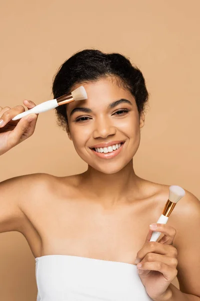 Femme afro-américaine souriante aux épaules nues tenant des pinceaux cosmétiques isolés sur beige — Photo de stock