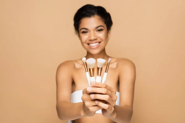 Verschwommen und fröhlich afrikanisch-amerikanische Frau mit nackten Schultern hält Kosmetikbürsten isoliert auf beige — Stockfoto