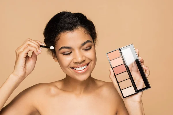 Happy african american woman with bare shoulders holding cosmetic brush and blush palette isolated on beige — Stock Photo