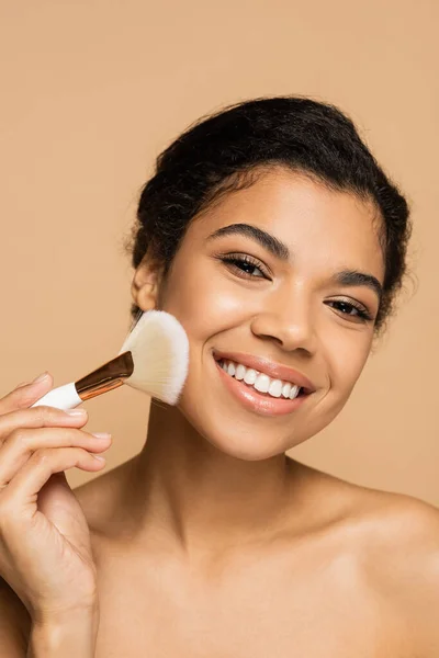 Smiling african american woman applying face powder with cosmetic brush isolated on beige — Stock Photo