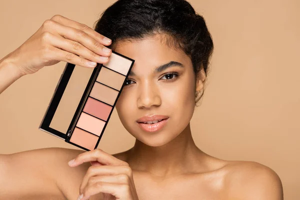 Brunette african american woman with bare shoulders holding cosmetic blush palette isolated on beige — Stock Photo
