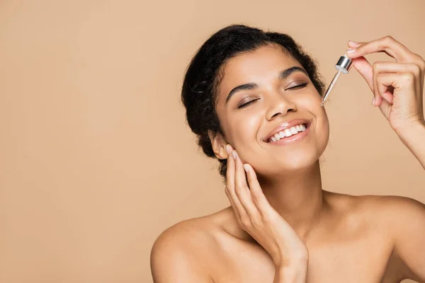 Happy african american woman with bare shoulders applying serum isolated on beige — Stock Photo