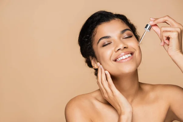 Mujer afroamericana feliz con hombros desnudos aplicando suero aislado en beige - foto de stock