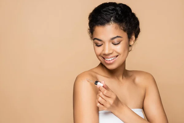 Happy african american woman with bare shoulders applying vitamin c serum on bare shoulder isolated on beige — Stock Photo