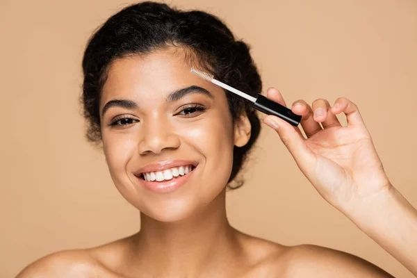 Smiling african american woman with bare shoulders styling eyebrow isolated on beige — Stock Photo