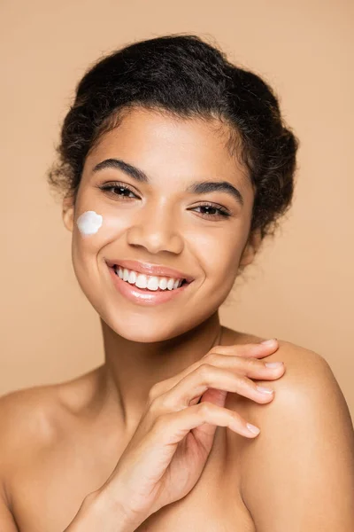 Joyful african american woman with face cream on cheek isolated on beige — Stock Photo