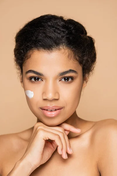 Brunette african american woman with face cream on cheek isolated on beige — Stock Photo