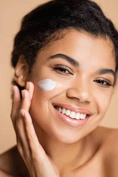 Souriant afro-américain femme appliquant crème visage sur la joue isolé sur beige — Photo de stock