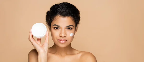 Morena afro-americana com creme facial em bochechas segurando recipiente isolado em bege, banner — Fotografia de Stock