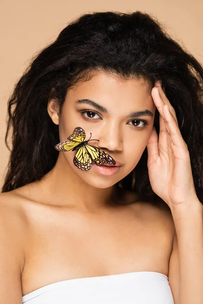 Jovem afro-americana com borboleta no rosto olhando para a câmera isolada no bege — Fotografia de Stock