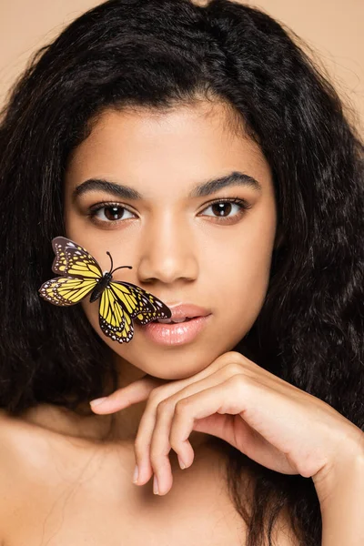 Brünette afrikanisch-amerikanische Frau mit dekorativem Schmetterling im Gesicht, der isoliert auf beige in die Kamera schaut — Stockfoto