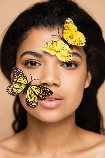 Jeune femme afro-américaine avec des papillons décoratifs sur le visage en regardant la caméra isolée sur beige — Photo de stock