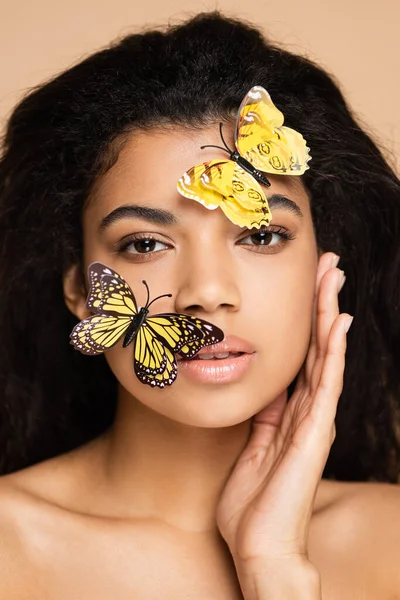 Брюнетка африканская американка с декоративными бабочками на лице, смотрящая на камеру, изолированную от бежевого — стоковое фото