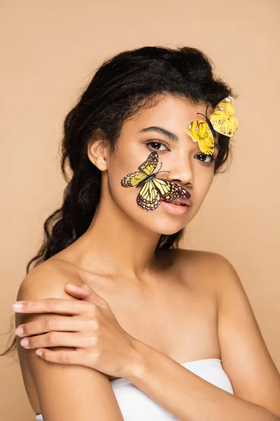 Morena mujer afroamericana con hombros desnudos y mariposas decorativas en la cara mirando a la cámara aislada en beige - foto de stock