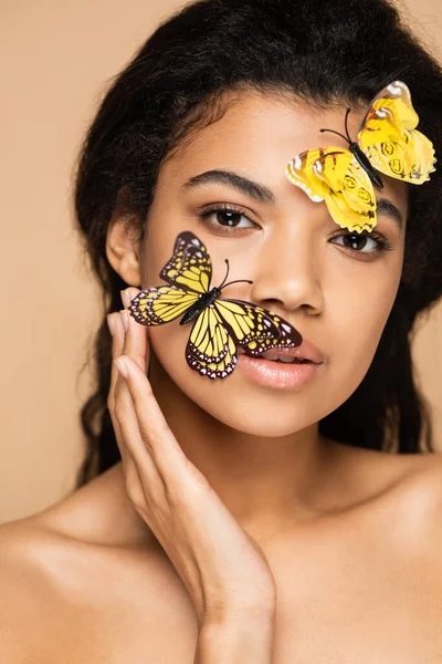 Jeune femme afro-américaine avec des papillons décoratifs jaunes sur le visage en regardant la caméra isolée sur beige — Photo de stock