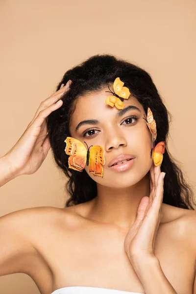 Joven mujer afroamericana con hombros desnudos y mariposas decorativas en la cara posando aislado en beige - foto de stock