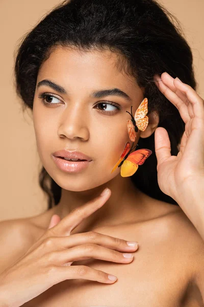 Joven mujer afroamericana con mariposas decorativas en la mejilla mirando hacia otro lado aislado en beige - foto de stock