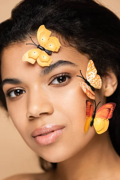 Primer plano de la joven afroamericana mujer con mariposas en la cara mirando a la cámara aislada en beige - foto de stock