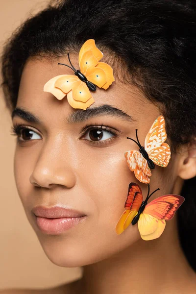 Gros plan de jeune femme afro-américaine avec des papillons sur le visage regardant loin isolé sur beige — Photo de stock