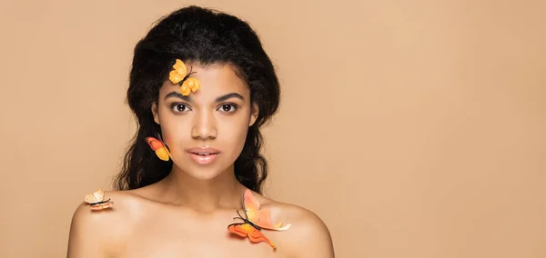 Mujer afroamericana bonita con mariposas anaranjadas en la cara y hombros desnudos aislados en beige, bandera - foto de stock