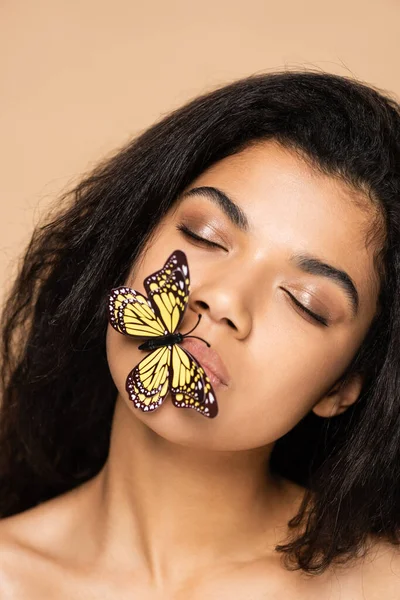 Mujer joven afroamericana con los ojos cerrados y la mariposa en los labios aislados en beige - foto de stock