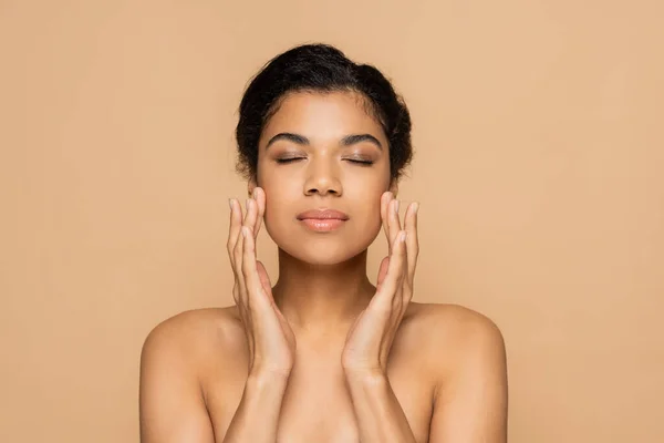 Mujer joven afroamericana con hombros desnudos y ojos cerrados tocando la cara aislada en beige - foto de stock