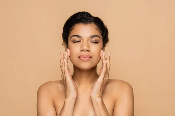 Mujer joven afroamericana con hombros desnudos y ojos cerrados tocando la cara aislada en beige — Stock Photo