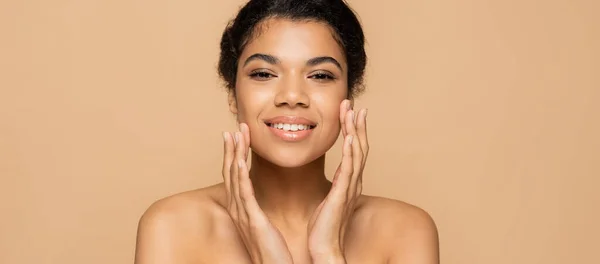 Happy african american woman with bare shoulders touching clean face isolated on beige, banner — Stock Photo