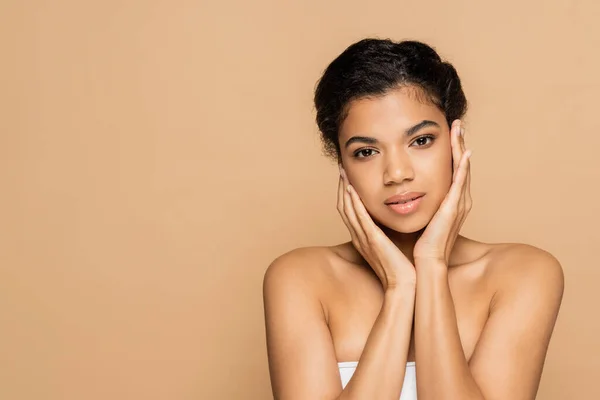 Young african american woman with bare shoulders touching clean face isolated on beige — Stock Photo