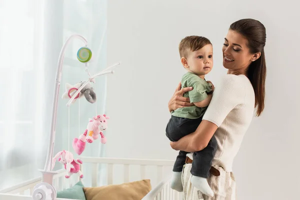 Mujer joven abrazando hijo cerca de cama de bebé con juguetes - foto de stock