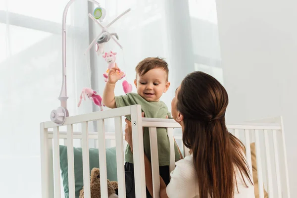 Frau hält fröhliches Kind neben Spielzeug im Babybett — Stockfoto