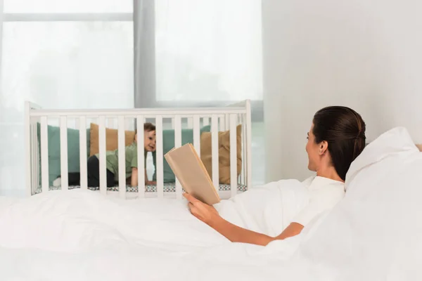 Frau mit Buch liegt auf Bett neben Kind im Babybett auf verschwommenem Hintergrund — Stockfoto