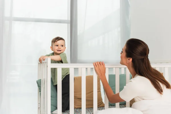 Smiling boy standing in baby bed near mother — Stock Photo
