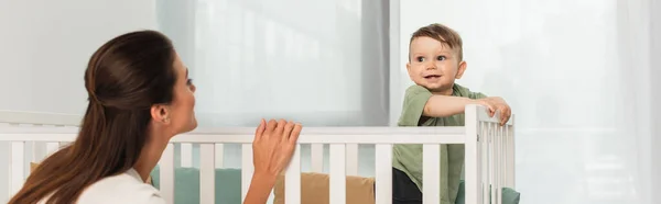 Femme debout près d'enfant souriant dans le lit de bébé, bannière — Photo de stock