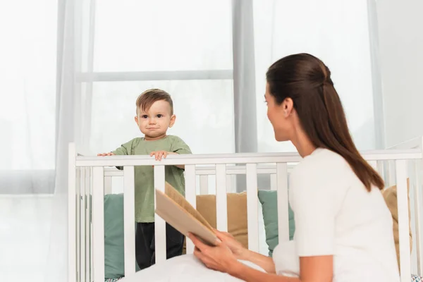 Kid standing in baby bed near blurred mother with book — Stock Photo
