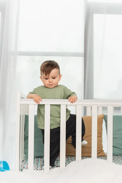 Niño pequeño de pie cerca de almohadas en la cama de bebé - foto de stock