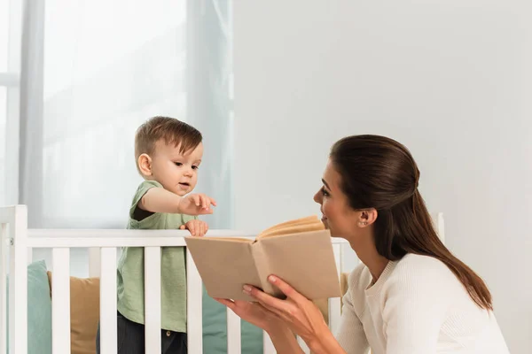 Smiling mother holding book near son in baby bed — Stock Photo
