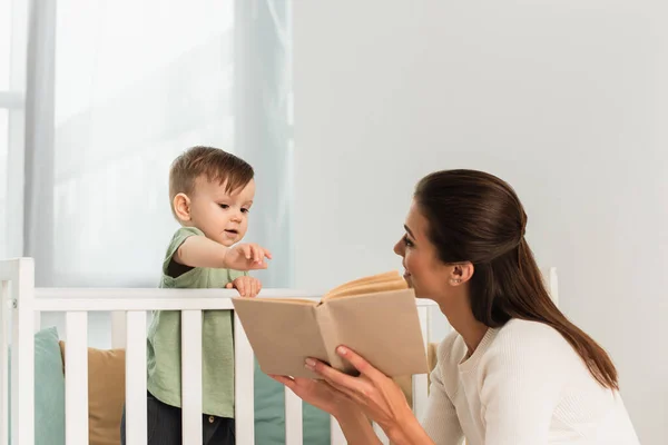 Sorrindo mãe segurando livro perto do filho na cama do bebê — Fotografia de Stock