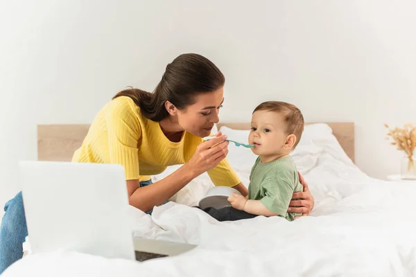 Mãe alimentando filho perto de laptop borrado na cama — Fotografia de Stock