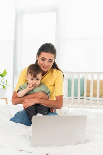 Frau umarmt Kleinkind neben verschwommenem Laptop im Schlafzimmer — Stockfoto