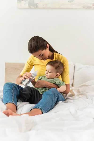 Young woman holding bottle of water near son on bed — Stock Photo