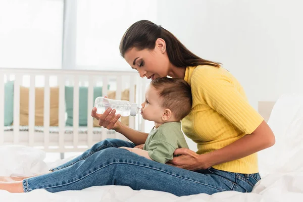 Vista lateral de la mujer sosteniendo botella de agua cerca de niño pequeño en la cama - foto de stock