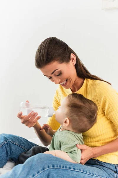 Smiling mother holding baby bottle while son drinking in bedroom — Stock Photo