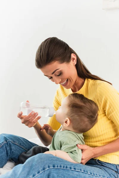 Sonriente madre sosteniendo biberón mientras su hijo bebe en el dormitorio - foto de stock