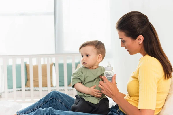 Woman holding baby bottle with pacifier near child on bed at home — Stock Photo