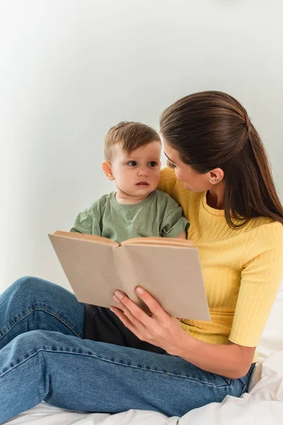 Young woman with blurred book looking at toddler son in bedroom — Stock Photo