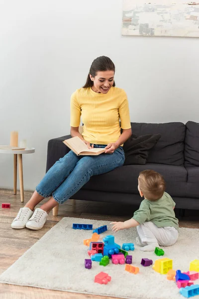 Happy mother holding book near kid with building blocks on carpet — Stock Photo