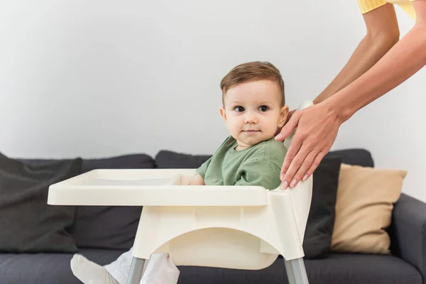 Madre de pie cerca de niño en la silla alta en casa - foto de stock